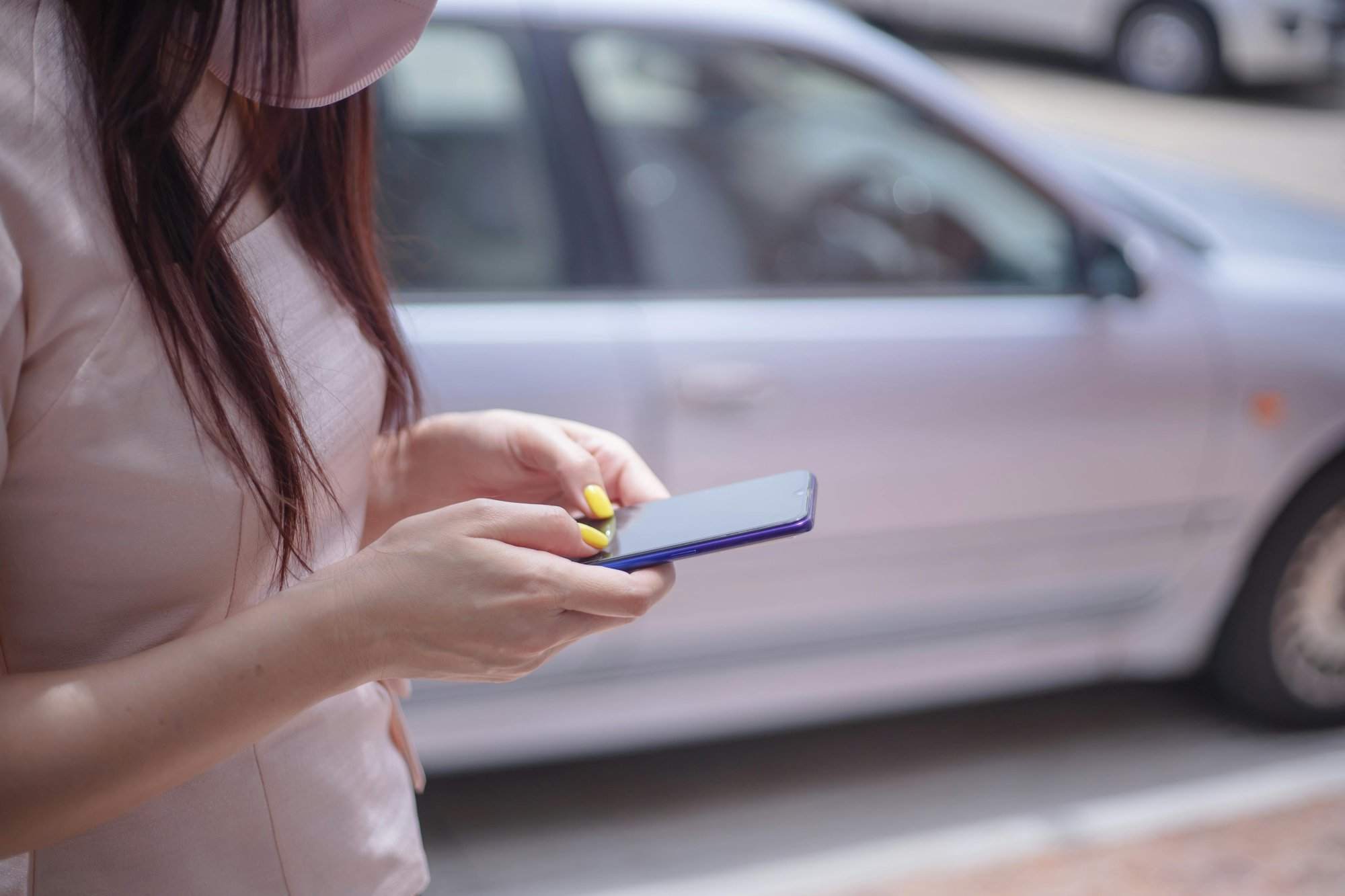 A close-up of a woman texting on a smartphone near a parked car outdoors.