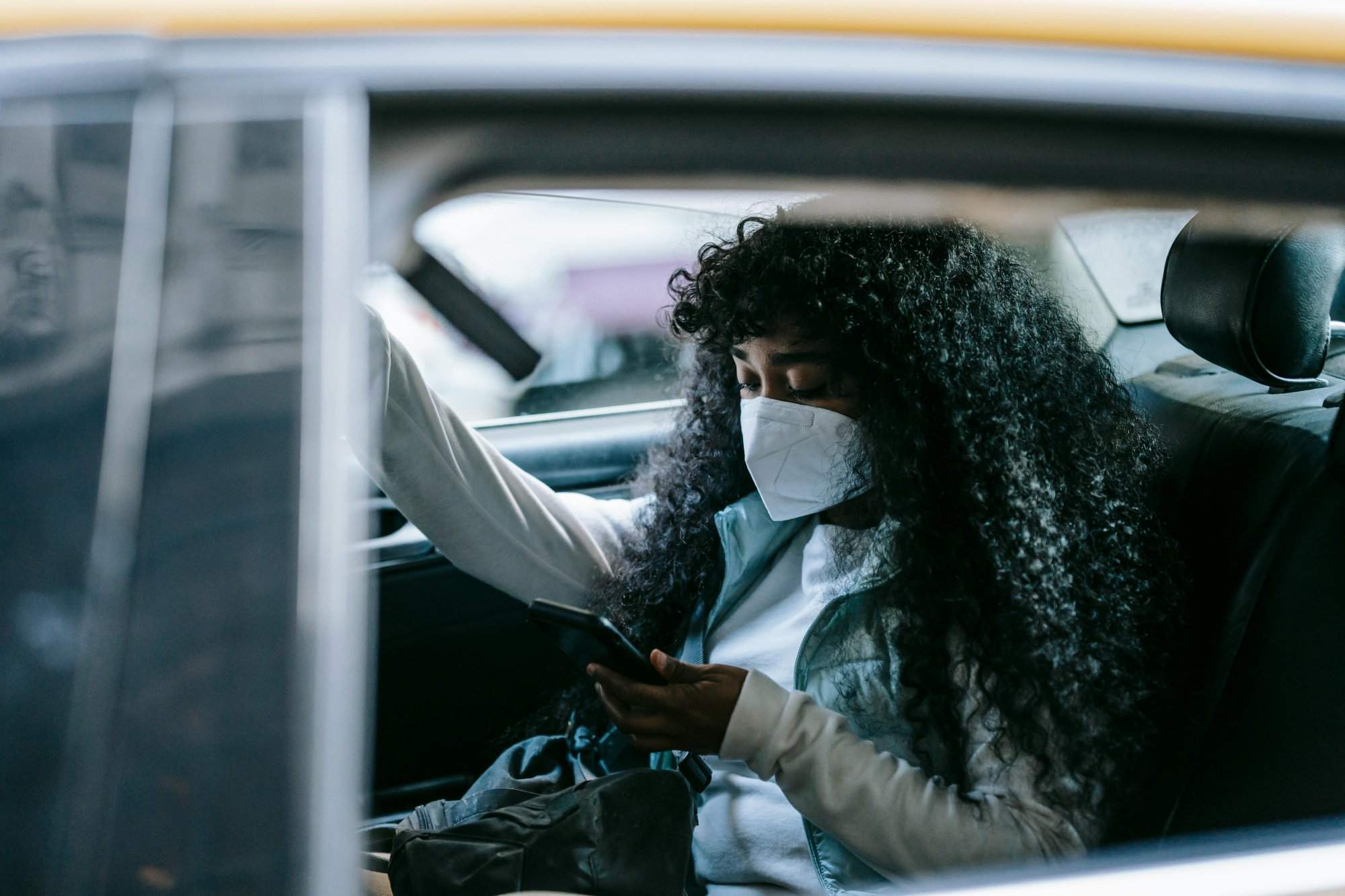 A woman with curly hair, wearing a mask, using her smartphone while commuting in a car.