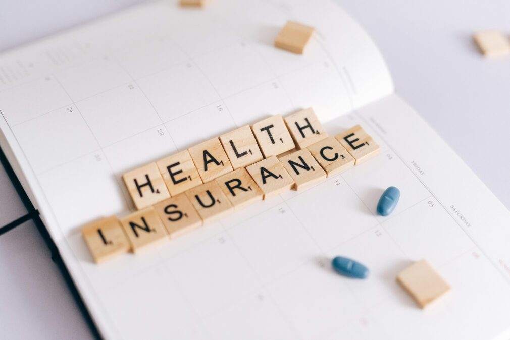 Close-up of health insurance spelled out with Scrabble tiles on a planner, alongside medication pills.
