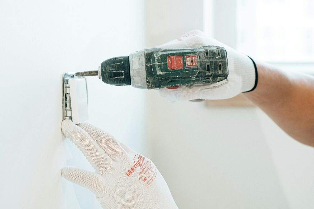 Gloved hands using a drill to install a power outlet on a wall, focusing on precision and safety.