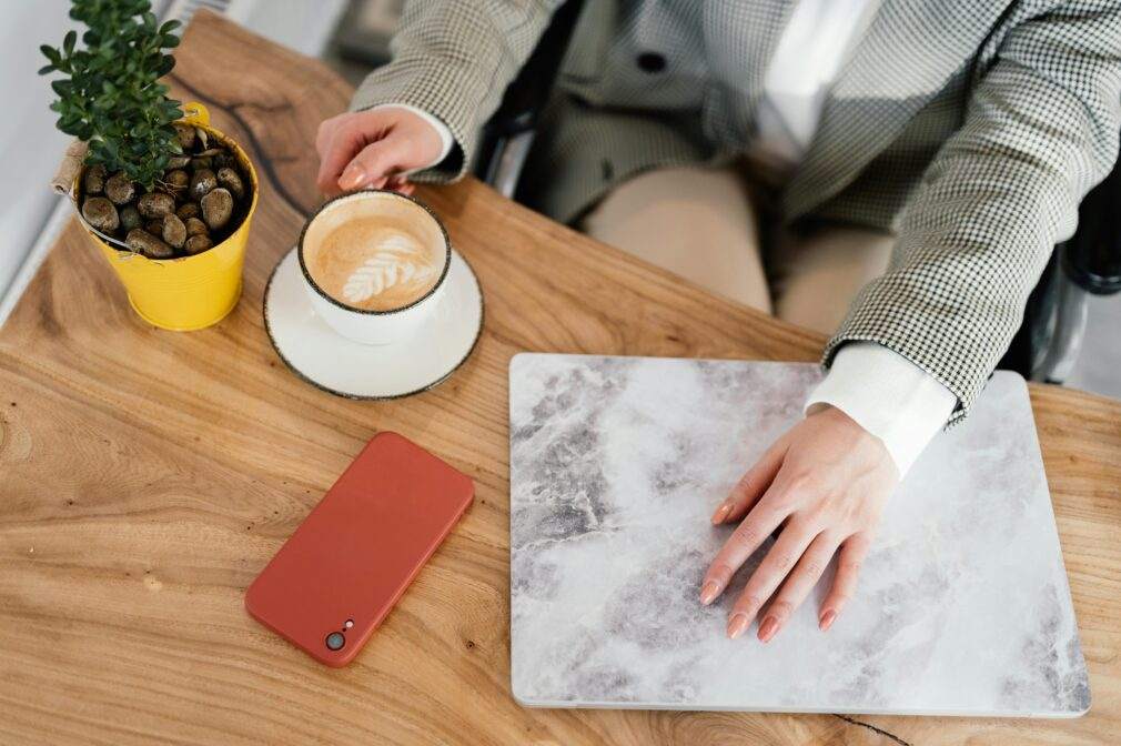 From above of unrecognizable female freelancer in wheelchair sitting at table with hot coffee and closed netbook during remote work in cafe