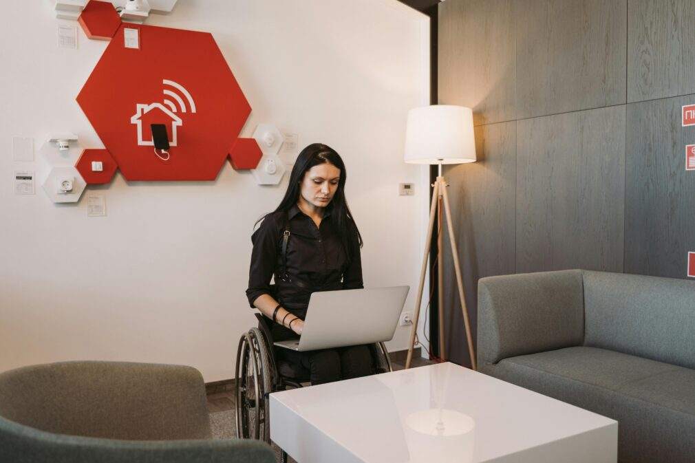 A woman in a wheelchair using a laptop in a modern office setting, highlighting accessibility and technology.