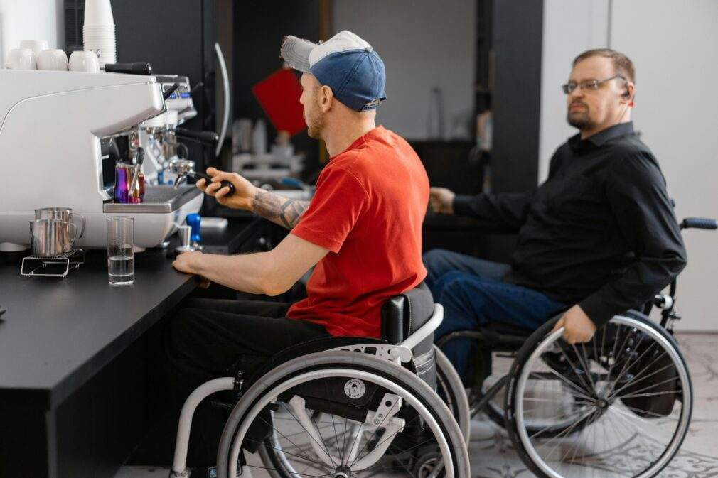 Two men in wheelchairs work at a coffee shop, showcasing inclusivity in the workplace.