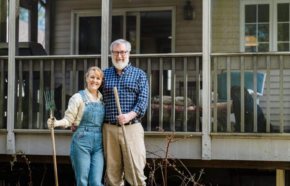 Elderly couple joyfully standing outside their home with gardening tools, symbolizing happiness and love.