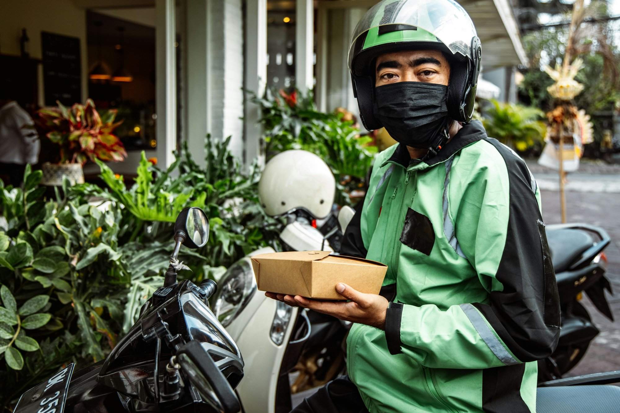 A delivery driver in green jacket holds food box while sitting on motorcycle.