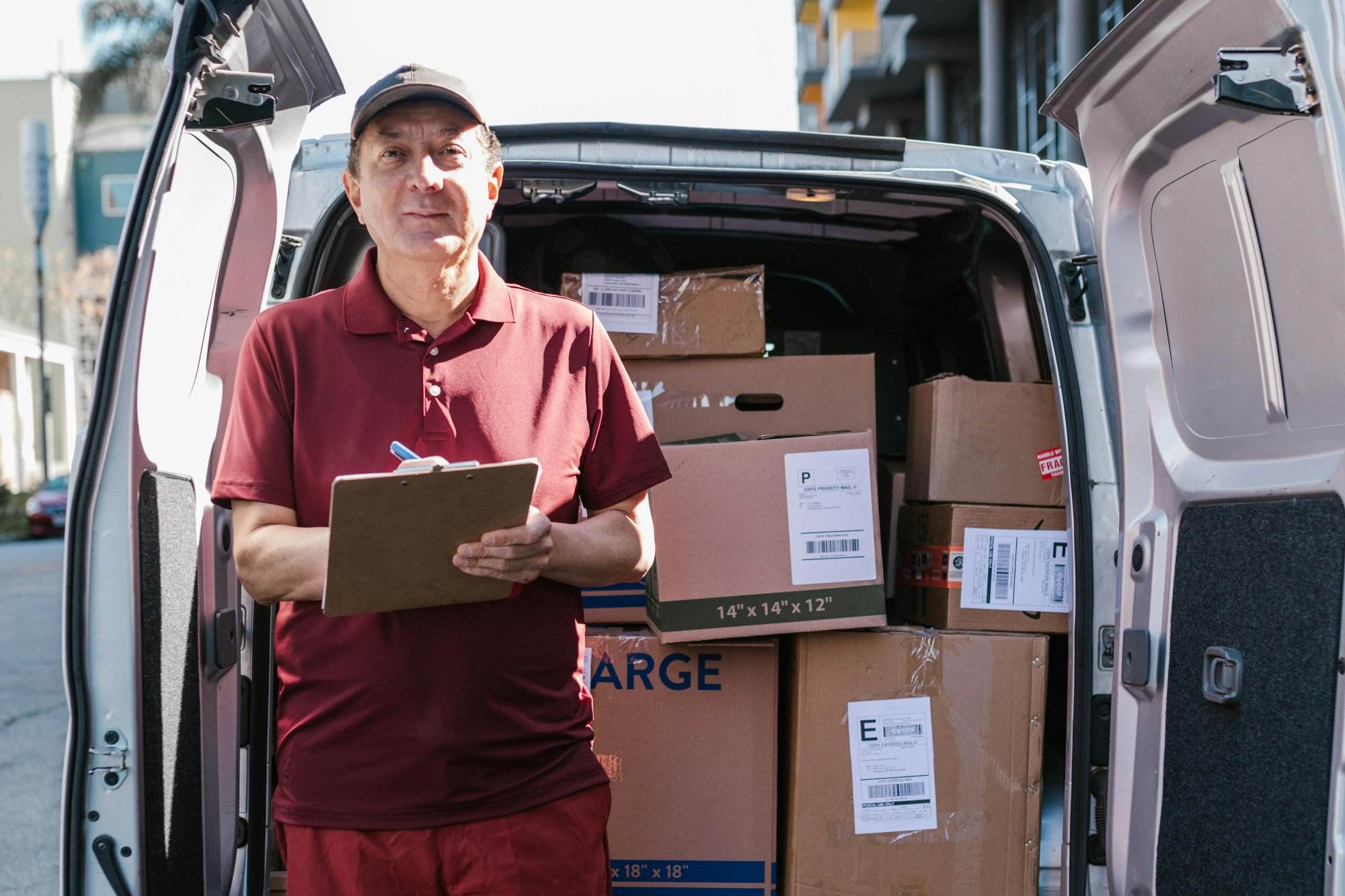 Elderly delivery person holds clipboard in front of open van filled with packages on a sunny day.