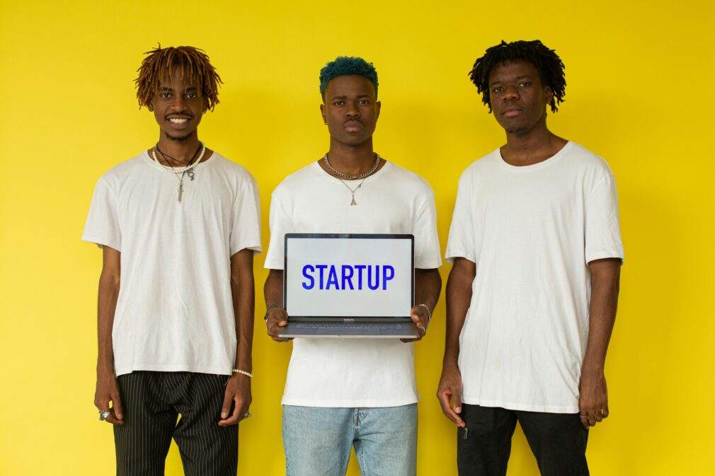 Three men holding a laptop displaying 'STARTUP' against a bold yellow background.