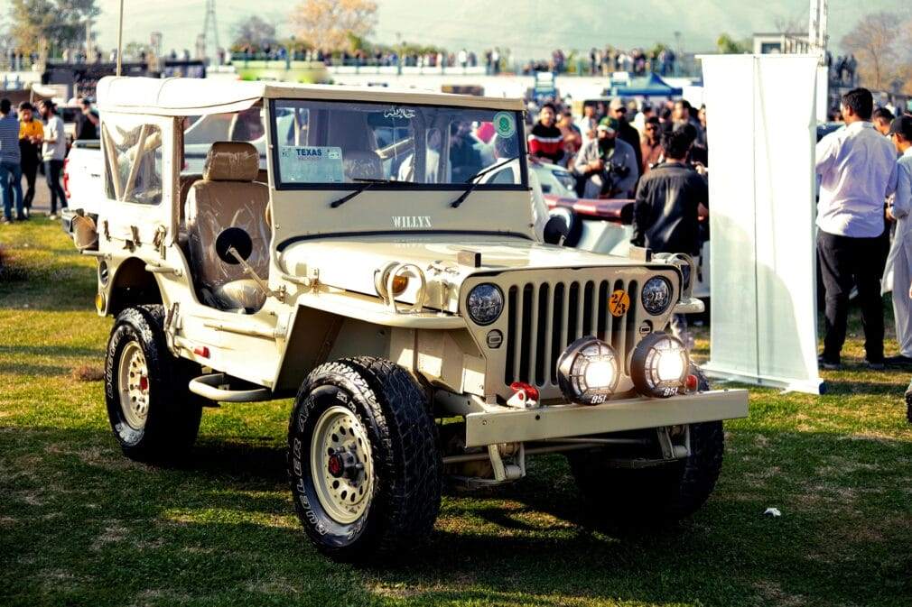 A classic Willys Jeep displayed at an outdoor car show, surrounded by people.