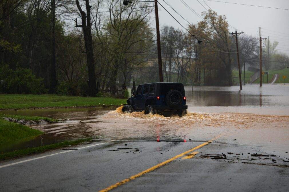 A jeep traverses a flooded road surrounded by trees, showcasing a challenging weather condition.