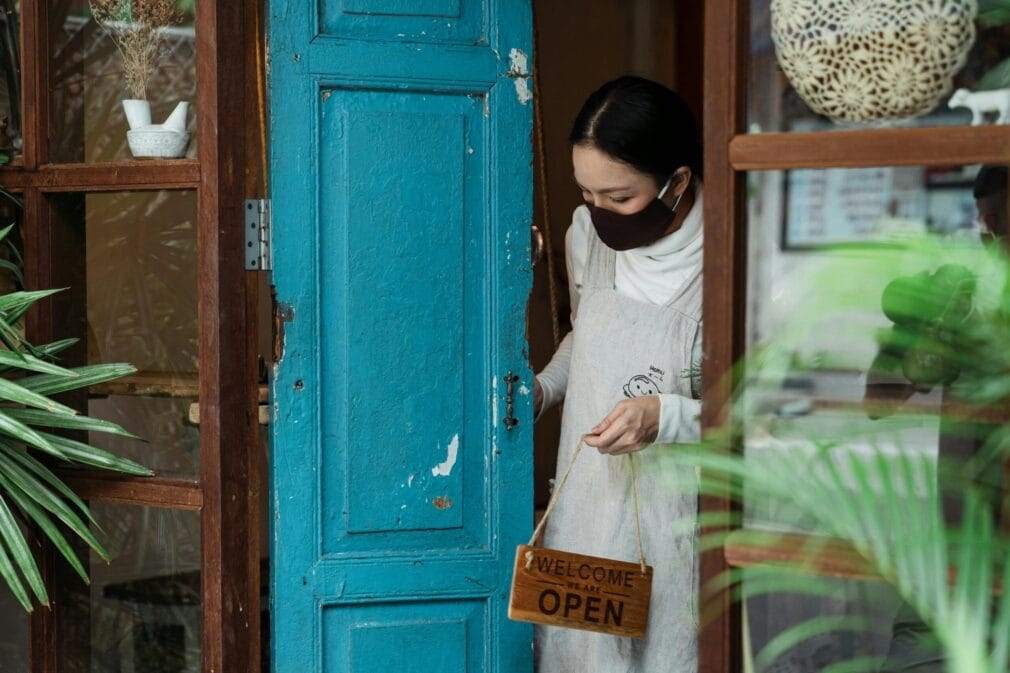 Woman wearing a mask opening the door of a vintage shop with a 'Welcome, We Are Open' sign.