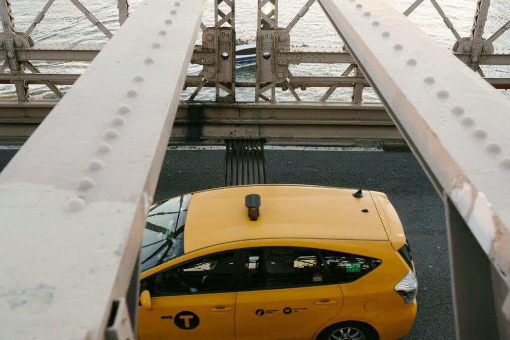 A New York City yellow taxi crossing the iconic Brooklyn Bridge over a river.
