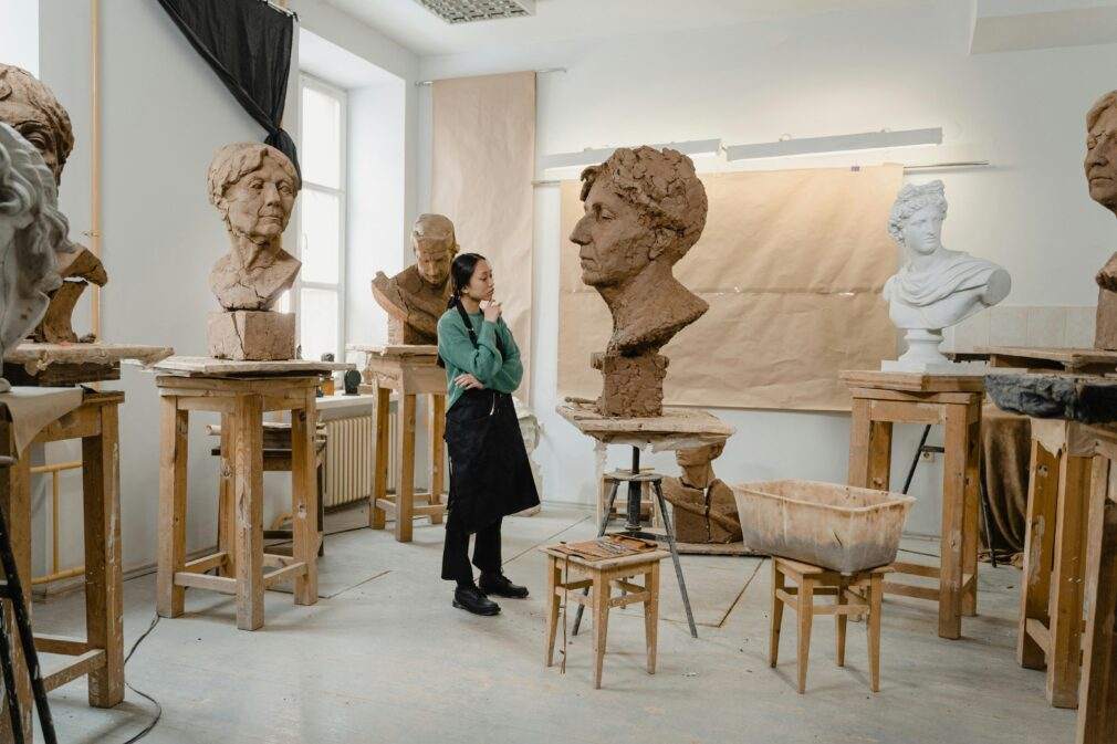 An artist in a creative clay sculpting studio surrounded by various sculpture busts on wooden stools.