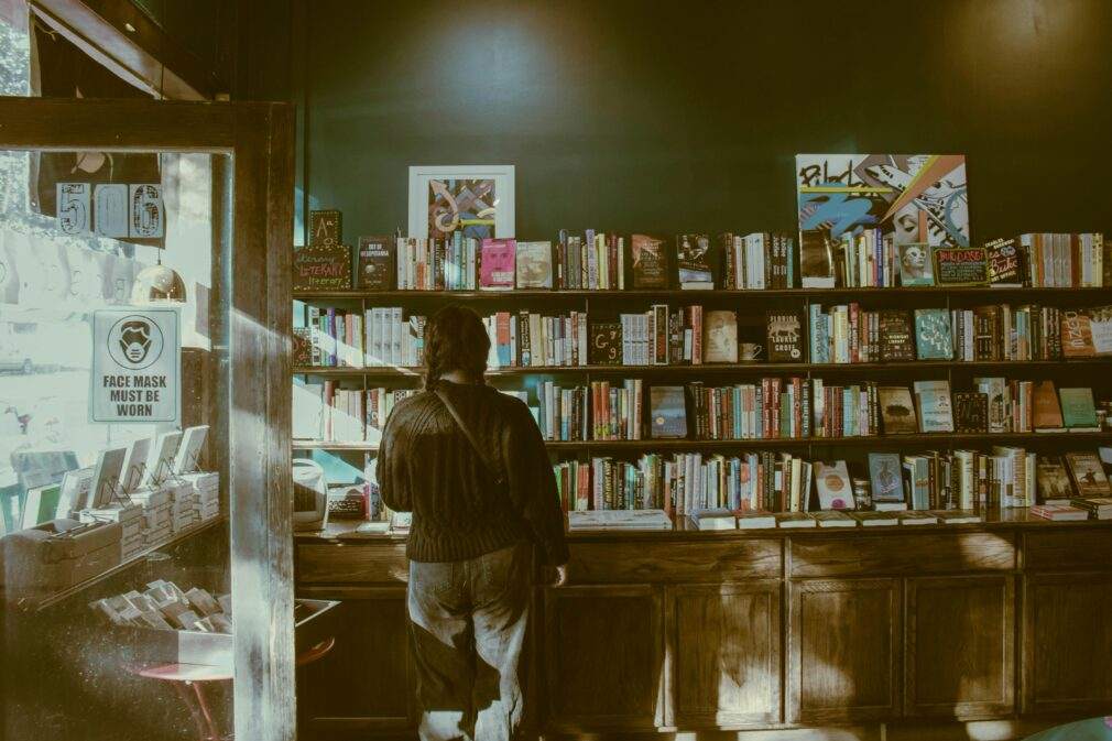 A person standing indoors, browsing books in a cozy bookstore with wooden shelves.