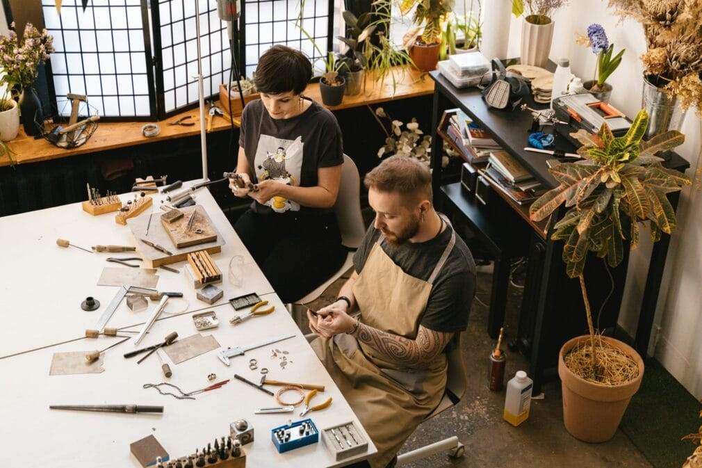 Two artisans working with tools and materials in a cozy, plant-filled workshop.