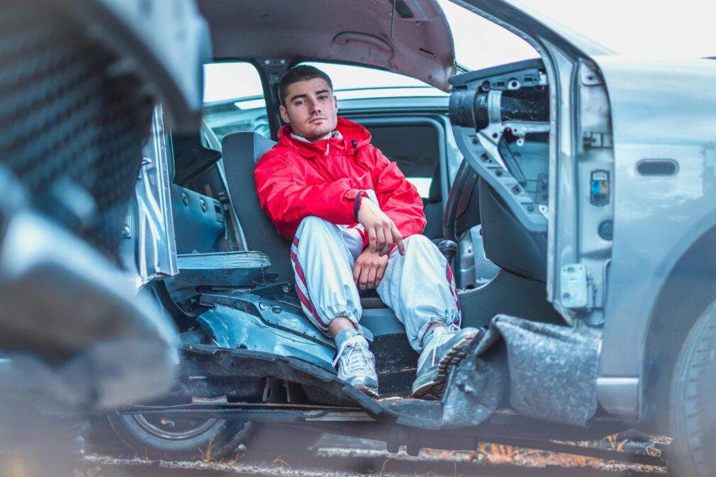 A young man in a vibrant red jacket sitting inside a damaged car, conveying themes of urban life and resilience.