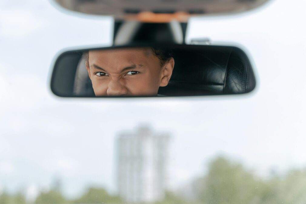A teenager practices driving, seen concentrating in a car's rearview mirror.